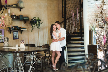 Young couple embracing in a cafe. A long legged shy girl smiling in her man's hands, wooden dark furniture around them
