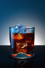 Whiskey glass with ice cubes on black glass surface over blue ba