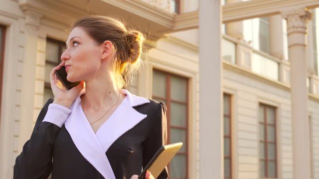 Smart elegant woman discussing business at mobile phone. She holding tablet and staying near office building. Close up shot with RL and LR panning