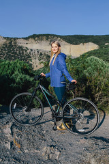 Obraz premium attractive young smiling blonde woman in a blue jacket standing with the bike and enjoying life the background mountains