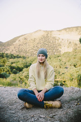 young blonde woman in a gray knitted hat, blue jeans and yellow working boots sitting outdoor on a background of mountains