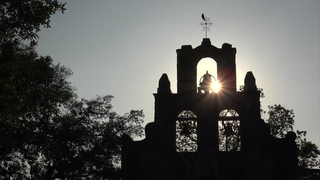 Spanish Mission Espada church bells in silhouette as bird lands on cross in San Antonio, Texas at sunset