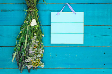 Blank wood sign hanging by bouquet of dried flowers with heart on rustic teal blue background