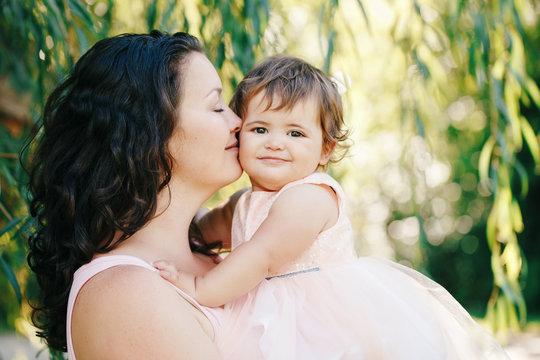 Lifestyle group portrait of beautiful white Caucasian brunette mother holding hugging daughter in pink dress kissing her in cheek on sunny spring summer day in park outside