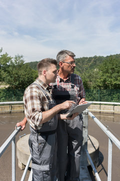 Two engineers standing on bridge over sedimentation tank monitoring the wastewater treatment with tablet computer