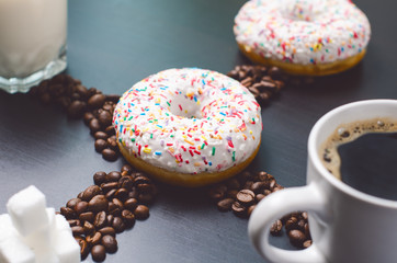 Donuts in a middle of picture. Coffee raw beans. Cup of coffee, sugar and milk. Breakfast on a dark blue table.