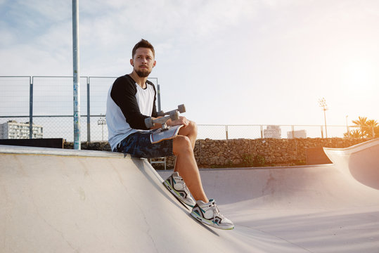 A brunet male in a blank white long sleeve t-shirt is sitting on a sunny background and looking aside. Young man holding skateboard is sitting in the urban park while relaxing.