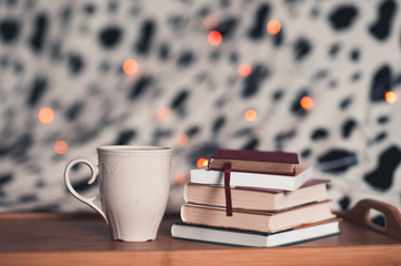 Cup of tea with stack of books stay on wooden tray in room over christmas lights.