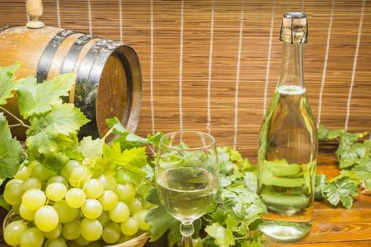 lambrusco,botella con vino blanco, copa de vino blanco, canasta con uvas blancas, hojas de vid alrededor y barril de vino al fondo de la imagen