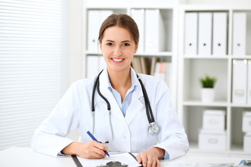 Young brunette female doctor sitting at the table and working at hospital office.  Health care, insurance and help concept. Physician ready to examine patient