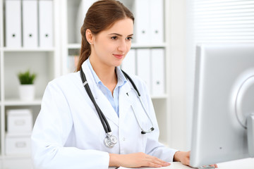 Young brunette female doctor sitting at the table and working at hospital office.  Health care, insurance and help concept. Physician ready to examine patient