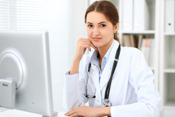 Young brunette female doctor sitting at the table and working at hospital office.  Health care, insurance and help concept. Physician ready to examine patient