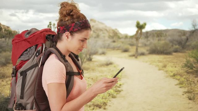 Cute woman hiker using smartphone to lookup the trail map through the desert. Side view of woman in her 20s using smartphone