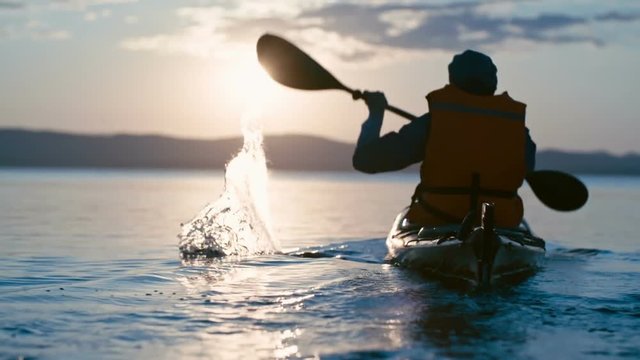 Slow motion rear view of male kayaker paddling away from camera towards sun setting above chain of mountains 