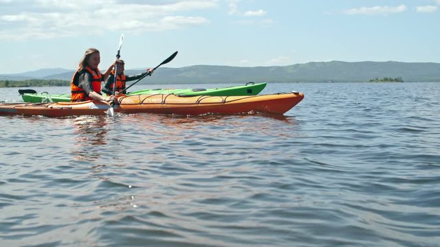 Slow motion surface level tracking of female and male athletes in life jackets paddling on touring kayaks with forward sweep strokes on blue water surface on sunny day 