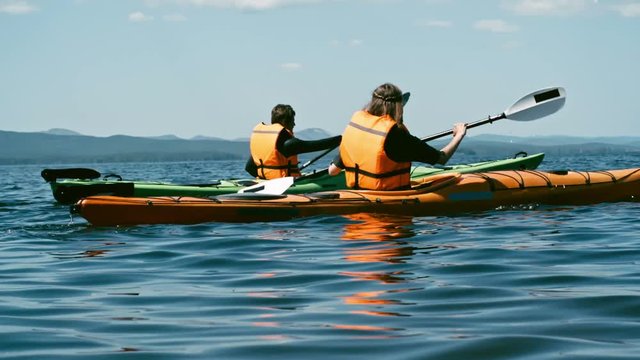 Slow motion rear view tracking of male and female athletes paddling kayaks along beautiful lake and looking at magnificent view of mountains 