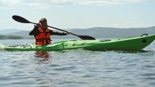 Slow motion side view tracking of male tourist in life jacket paddling touring kayak across blue lake with picturesque chain of mountains visible in background