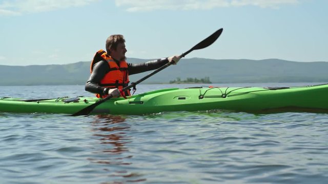 Slow motion tracking of man with beard forward sweeping on his kayak across blue lake 