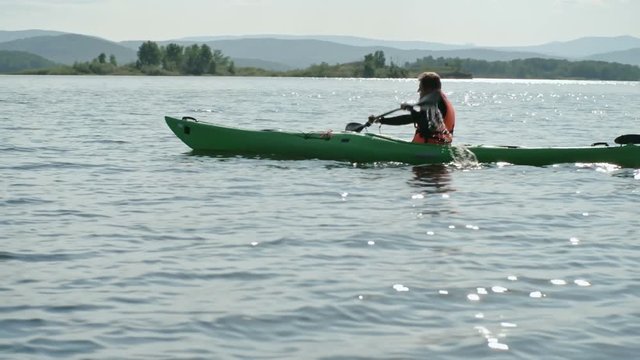 Slow motion tracking of male tourist kayaking on blue lake water with forward sweep strokes, mountains visible in distance 