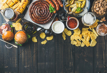Beer and snack set. Octoberfest food frame concept. Variety of beers, grilled sausages, burgers, corn, fried potatoes, chips, salted almonds and sauces on dark wooden background. Top view, copy space