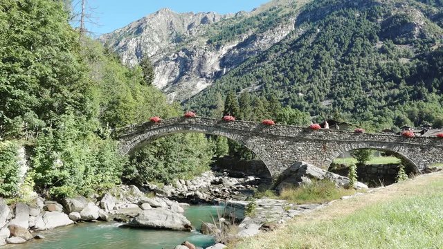 Gaby, Aosta valley, Italy. People walking on old medieval bridge with mountains landscape, Lys river and flowers on the ancient path. Summer holidays, nature and italian tourism