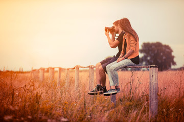 Young couple sitting on the fence in the summer sunlight and taking pictures with a digital camera, friendship and relationships concept