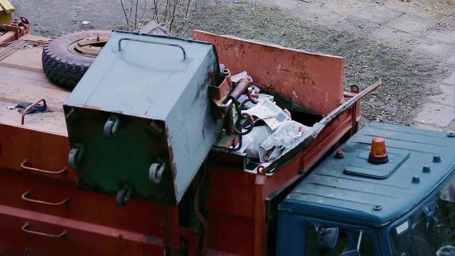 Garbage truck sweeps the contents of waste container into the hopper. Orange-blue truck with emergency vehicle lighting.
