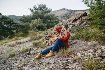 portrait of a beautiful young blonde woman in hat, red sweater, blue jeans and yellow boots sits outdoor on the background of mountain and forest