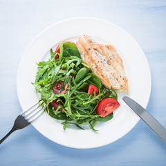 Roasted chicken breast and fresh salad with tomato and greens (spinach, arugula) top view on blue wooden background. Healthy food.