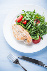 Roasted chicken breast and fresh salad with tomato and greens (spinach, arugula) top view on blue wooden background. Healthy food.