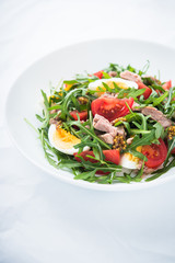 Fresh salad with tuna, tomatoes, eggs, arugula and mustard on white textured background close up. Healthy food.