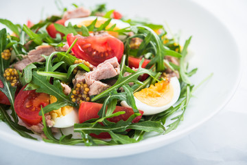Fresh salad with tuna, tomatoes, eggs, arugula and mustard on white textured background close up. Healthy food.
