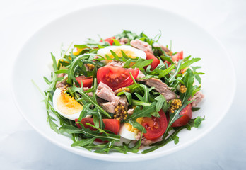 Fresh salad with tuna, tomatoes, eggs, arugula and mustard on white textured background close up. Healthy food.