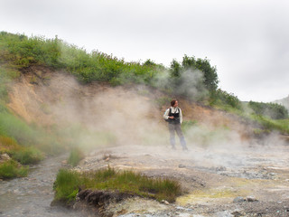 Adult woman with a backpack standing in a smoking crater of the volcano Mutnovsky on Kamchatka in Russia against the background of a hill with trees and sky with clouds