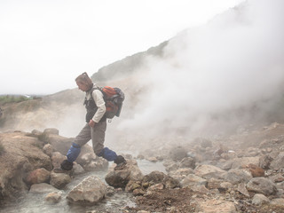 Adult woman with a backpack jumping over a hot stream in a smoking crater of the volcano Mutnovsky on Kamchatka in Russia against the background of a hill with trees and sky with clouds