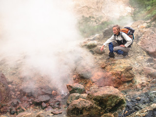 Adult woman with a backpack sitting in a geyser on smoking crater of the volcano Mutnovsky on Kamchatka in Russia against the background of a hill with grass