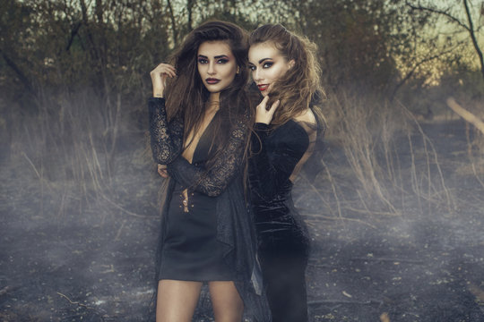 Two young beautiful sexy witches in black gowns standing in the middle of burnt meadow with predatory face expression. Halloween concept. Outdoors shot.