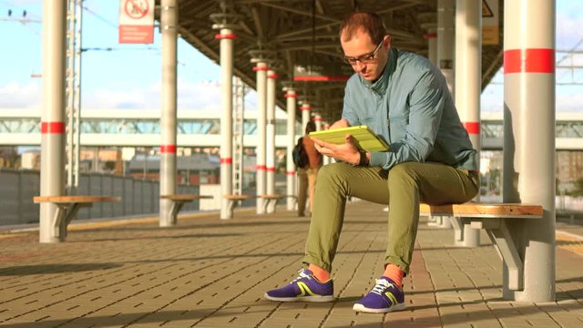 Bald nerdy man in glasses sitting at railroad station and using his tablet computer at sunset. 4K shot