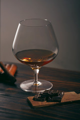 Glass of brandy on a wooden table with chocolate and cigar. gray background.