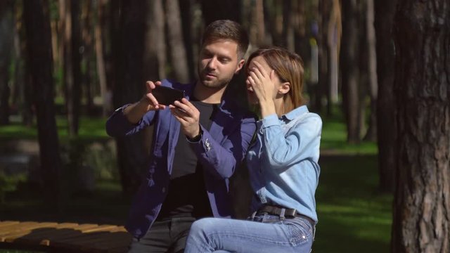 Happy pair in love take selfie photo on the bench in park. Caucasian woman and handsome man photographed have fun outdoors on a date. Guy using smartphone girlfriend make funny pose.