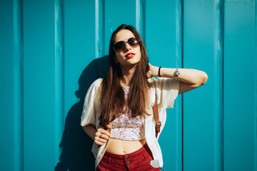 A beautiful model look girl with long brown hair wearing casual clothes is standing beside the wall. A caucasian brunette female wearing sunglasses is posing on the light blue background.