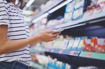 Woman shopping healthy food in supermarket background. Close up view girl buy products using digital gadget in store. Hipster at grocery using smartphone. Person comparing the price of produce