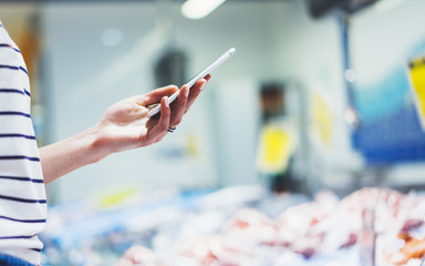 Woman shopping healthy sea food in supermarket background. Close up view girl buy products using digital gadget in store. Hipster at grocery using smartphone. Person comparing the price of produce