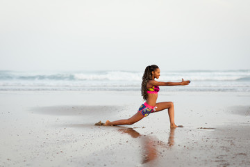 African woman stretching legs in yoga pose at the beach. Sporty fit female dancer exercising against the sea.