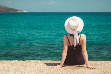 Fototapeta na wymiar Rear view of stylish female wearing sun hat, sitting on edge of concrete dock and enjoying the sea view. Travel and summer vacation concepts. 