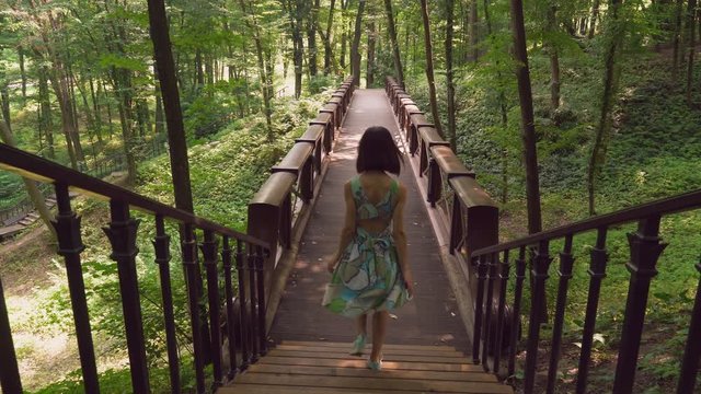 Young brunette woman in a bright dress down downstairs and walking on the bridge in the park on a sunny day. Rear back view