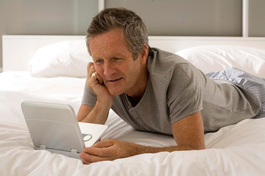 Puzzled senior man lying on bed staring at laptop