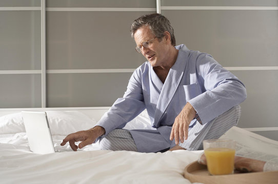 Puzzled senior man on bed typing on laptop