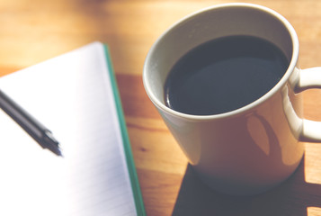 A cup of black coffee and an open notebook. A pencil is also on the wooden table. Image has a vintage effect.