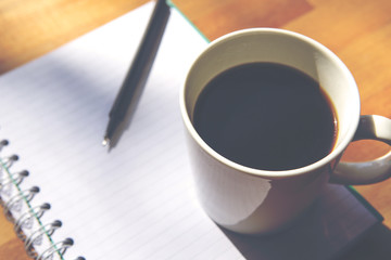 A cup of black coffee and an open notebook. A pencil is also on the wooden table. Image has a vintage effect.